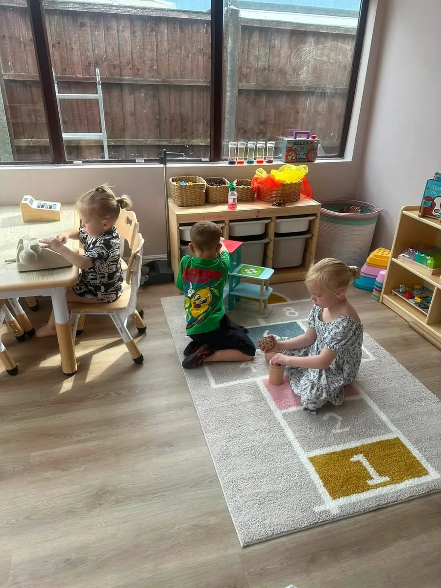 Three children playing in the bright playroom on a hopscotch rug, with wooden shelves and baskets of toys