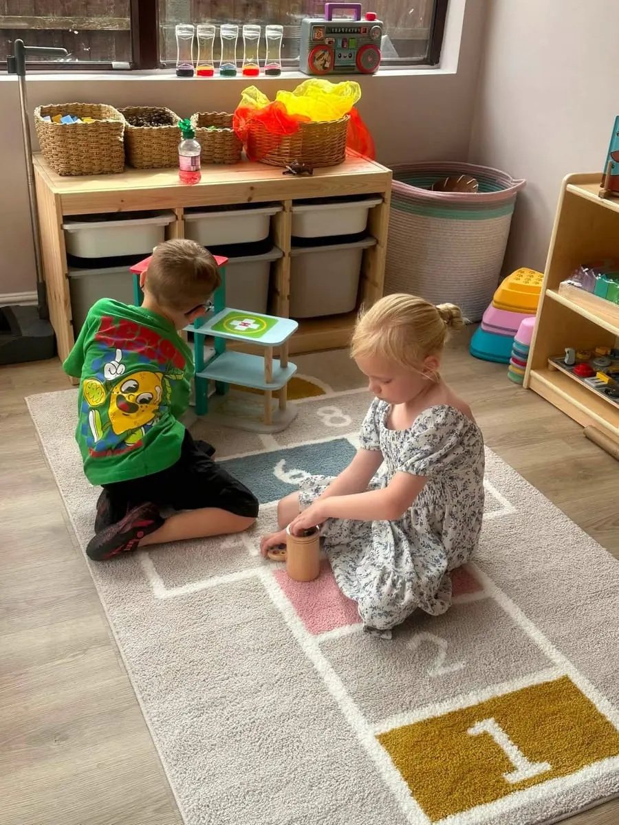 Two children sitting together on the playroom rug, exploring wooden toys side by side