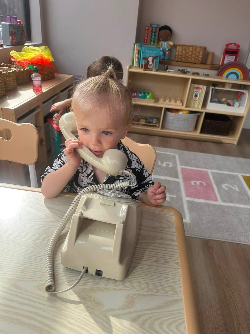 A toddler chatting on a toy telephone at a low wooden table in the playroom